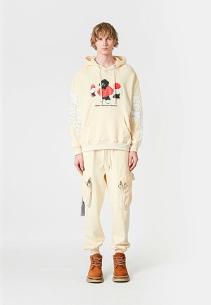 Joven vestido con una sudadera beige con un gráfico de dibujos animados y pantalones cargo a juego, de pie contra un fondo blanco liso.