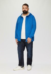 Niebieska rozpinana bluza z szarym wnętrzem, z dwiema przednimi kieszeniami, w połączeniu z ciemnymi dżinsami i białymi trampkami. Prosty, casualowy design.