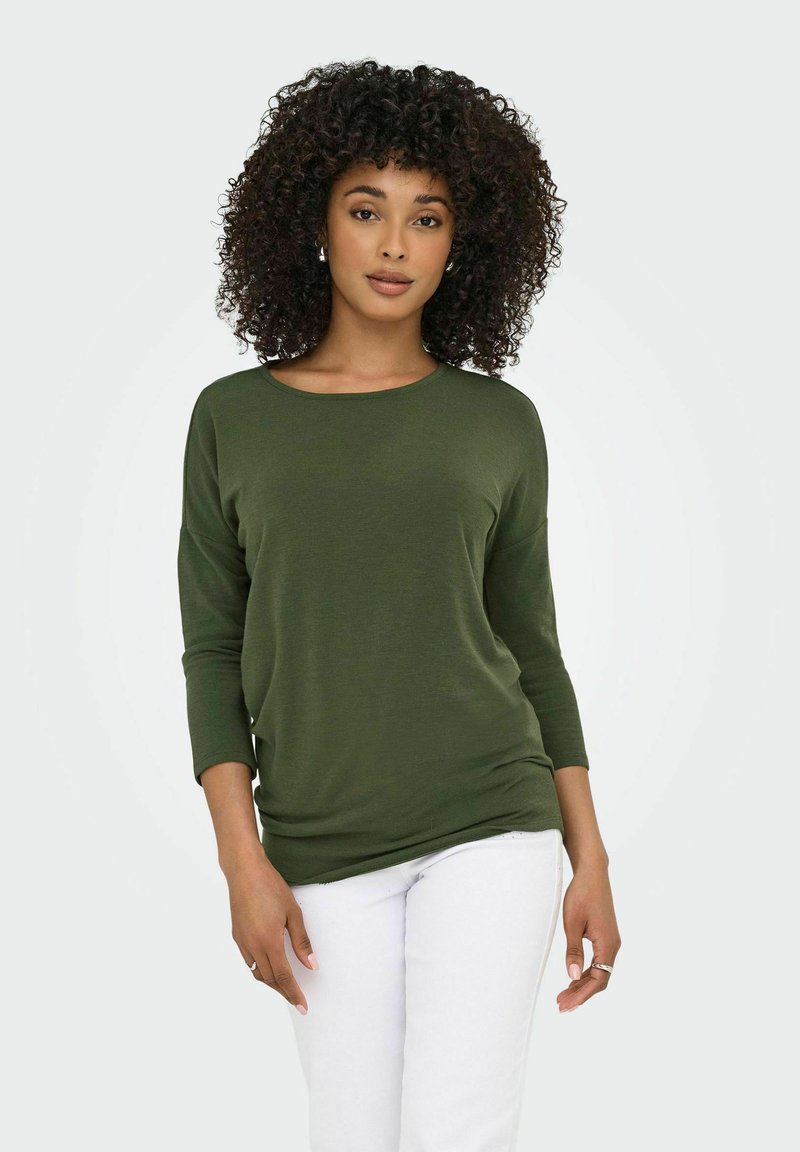 Top verde oliva a maniche lunghe con scollatura rotonda e vestibilità ampia, realizzato in tessuto morbido. Abbinato a pantaloni bianchi.