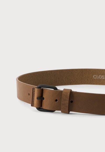 Ceinture en cuir marron avec boucle et passant en métal noir sur fond blanc, partiellement enroulée pour montrer la texture et les coutures.