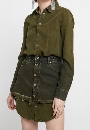 Nainen, joka pukeutunut oliivinvihreään kaulukselliseen paitamekkoon, jota on kerroksellisesti yhdistetty tummanvihreään farkkujen lyhythihaiseen minihameeseen, jossa on repsottava helma.