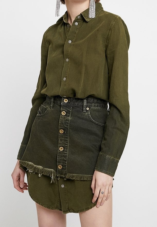Femme portant une robe chemise boutonnée vert olive superposée à une mini-jupe en jean vert foncé à bout effiloché.