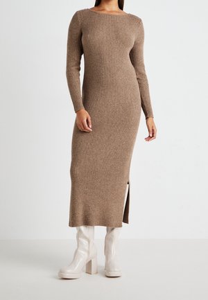 Frau trägt ein langes, geripptes, braunes Strickkleid mit langen Ärmeln und einem Seitenschlitz, kombiniert mit weißen Stiefeletten mit Blockabsatz.