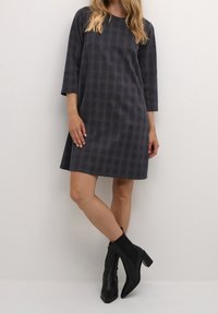 Vestito a scacchi grigio scuro con maniche a tre quarti, vestibilità ampia, orlo uniforme. Indossato con stivaletti neri alla caviglia con tacco a blocco.