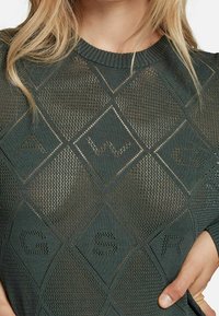 Suéter de punto en verde oscuro con un patrón de diamantes. Presenta un cuello acanalado y una textura en todo su diseño, destacando las letras A, W, G, S, R.