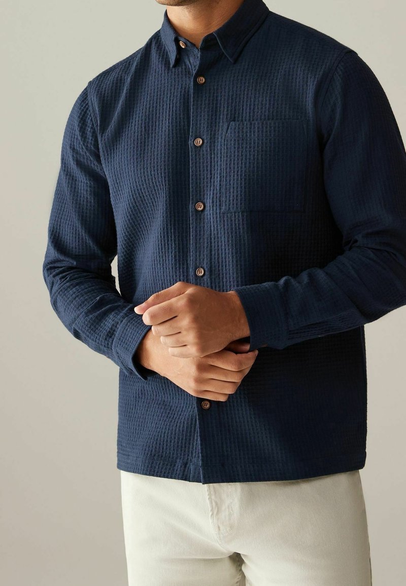 Homme portant une chemise boutonnée bleu marine texturée avec une poche avant, poignets à bouton, associée à un pantalon beige clair.