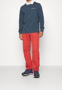 Marineblauwe polo met lange mouwen en een witte kraag, gecombineerd met rode broek en marineblauwe wandelschoenen. De polo heeft een klein logo op de borst.