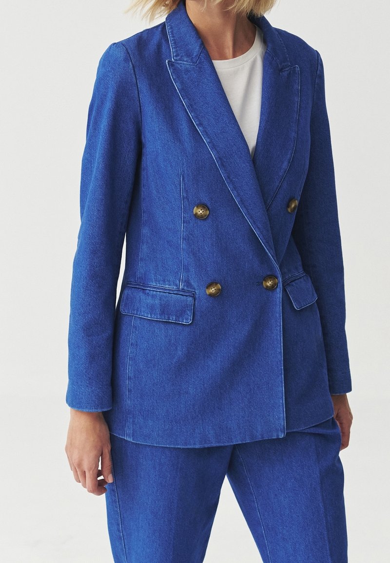 Femme portant un blazer bleu croisé avec des boutons dorés et un pantalon assorti par-dessus un top blanc.