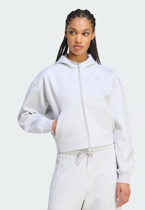 Ung kvinde med fletninger iført en lysegrå hættetrøje med lynlås og matchende joggers, der kigger til venstre med hånden i en lomme.