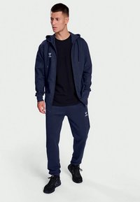 Haut de sport à capuche bleu marine avec fermeture éclair, capuche ajustable et logo, associé à un jogging assorti. Les deux sont fabriqués dans un tissu doux et texturé.