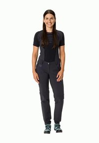 Γυναίκα που στέκεται και χαμογελάει, φορώντας ένα μαύρο κοντομάνικο μπλουζάκι, σκούρες cargo παντελόνες και teal ορειβατικά παπούτσια, σε λευκό φόντο.