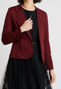 Burgundy istuva bleiseri, jossa on lovettu kaulus, kaksi nappia ja kiinteä kangas. Päällä musta paita, yhdistettynä puolisiirtoon mustaan hameeseen.