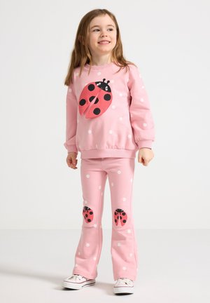 Jong meisje dat een roze trui met witte stippen en een broek met grote lieveheersbeestjes draagt, glimlachend en staand tegen een effen achtergrond.