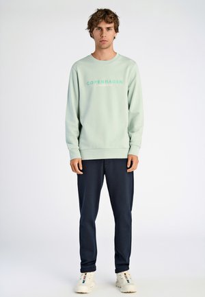 Lys grøn sweatshirt med "COPENHAGEN" tekst, sammen med mørk marineblå bukser, iført hvide sneakers. Glat stof, afslappet design.