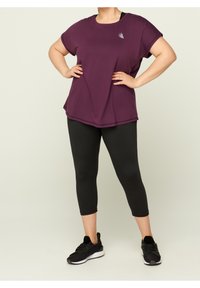Fioletowa, krótki rękaw sportowa bluzka o delikatnym kroju A, zestawiona z czarnymi leginsami 3/4 i czarnymi butami sportowymi.