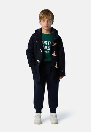 Cappotto navy con bottoni a fibbia in legno, felpa verde con grafica, pantaloni jogger navy e sneakers bianche. Outfit casual a strati per bambini.