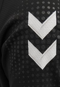 Sort tekstureret stof med et prikket mønster. Har to hvide chevron-designs med bølgelinjer, placeret lodret på siden.