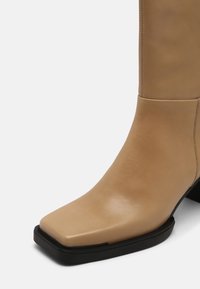 Beżowy skórzany botek za kostkę z kwadratowym noskiem, gładką fakturą i czarnym, prostokątnym obcasem. Czyste linie z minimalnymi detalami przeszyć.