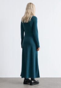 Frau mit langen blonden Haaren, die ein enganliegendes türkisfarbenes Rippenstrickkleid und schwarze Slipper trägt, steht gegen eine schlichte weiße Wand.