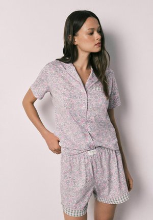 Jonge vrouw draagt een lichtroze bloemenprint kort mouw knoop top met bijpassende shorts met geruite rand, staand met hand op heup.