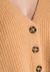 Sweter kardigan z dekoltem w serek w miękkim, brzoskwiniowym kolorze. Posiada prążkowaną teksturę oraz cztery okrągłe, ciemne guziki z żółwiowej skorupy.