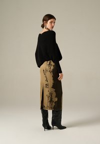 Femme portant un pull noir à manches longues, une jupe midi beige à fleurs avec une fente, et des bottes noires à talons hauts, se tenant devant un fond uni.