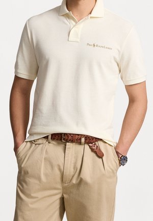 Homme portant un polo Ralph Lauren crème à manches courtes, un pantalon plissé beige, une ceinture en cuir tressé marron et une montre-bracelet à cadran sombre.
