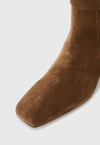 Bota de tornozelo em camurça castanha com biqueira quadrada, textura suave e detalhes de costura mínimos, apresentando um salto alto e uma silhueta elegante.