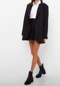 Czarna, oversize marynarka, biała koszula z kołnierzykiem, czarna plisowana mini spódniczka oraz czarne botki na obcasie z teksturowaną podeszwą.