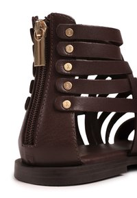 Sandale plate marron foncé avec plusieurs brides horizontales, fermeture éclair dorée au talon, et petits clous ronds dorés aux extrémités des brides.
