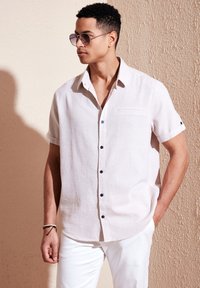 Camisa de botón de manga corta en color beige claro con un bolsillo en el pecho y botones oscuros, combinada con pantalones blancos y gafas de sol redondas.