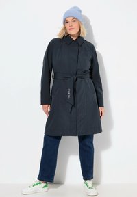 Cappotto trench blu scuro, lungo fino alla vita, con cintura, maniche lunghe e tasche laterali, indossato sopra jeans scuri e sneakers bianche. Berretto azzurro.