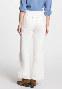 Weiße weite Baumwollhosen mit aufgesetzten Taschen und subtilen Steppdetails, kombiniert mit braunen Wildleder-Stiefeln bis zu den Knöcheln.