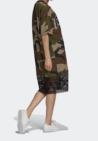 Camo-patterned oversized t-shirt dress with a black lace hem, featuring short sleeves and a rounded neckline. Paired with white sneakers.