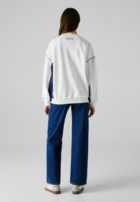 Felpa bianca con accenti multicolore, polsini e orlo a costine. Abbinata a jeans a gamba larga blu e sneakers bianche. Dettaglio logo semplice sul retro.
