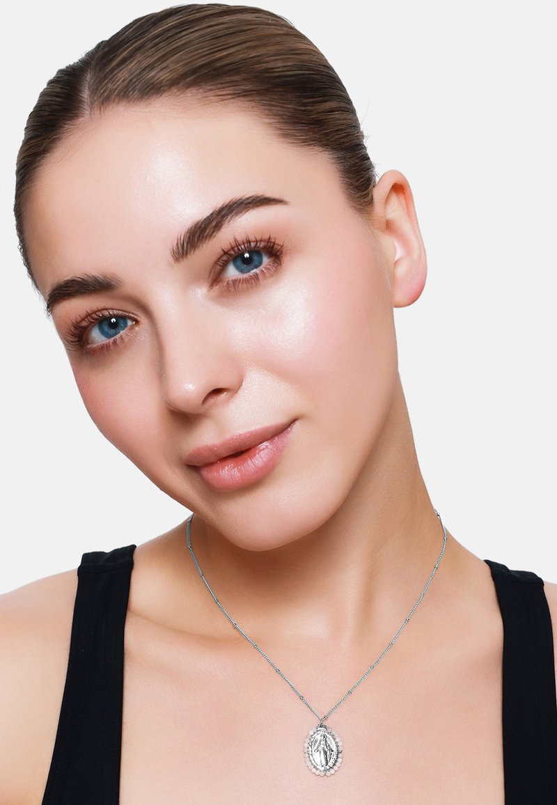 Collier en argent avec un pendentif ovale présentant des détails complexes, porté par une personne aux cheveux bruns raides, portant un haut noir sur fond blanc.