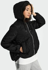 Schwarze gesteppte Steppjacke mit Kapuze, ausgestattet mit einem Frontreißverschluss und Kordelzugdetails. Akzente setzen elastische Bündchen und eine lockere Passform.
