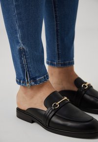 Svarta läderskor utan snörning med en detalj i guld, kombinerade med blå denimjeans med dragkedjor vid anklarna. Lätt struktur synlig.