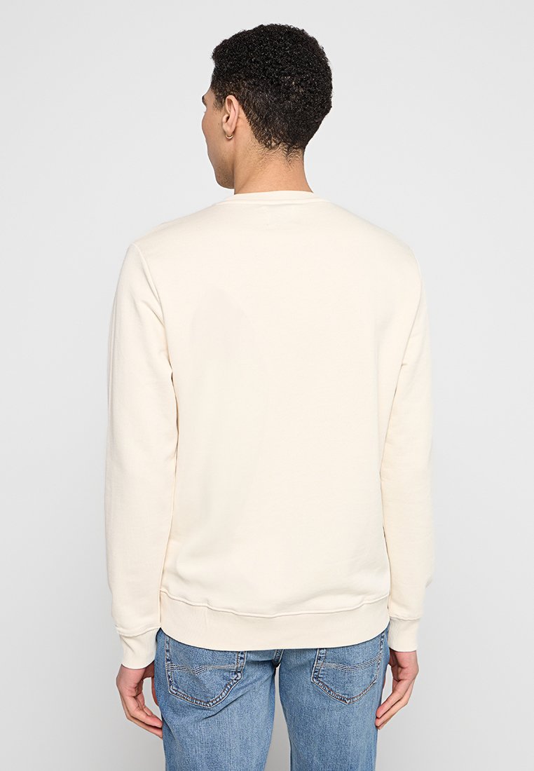 Cremefarbener Sweatshirt mit Rundhalsausschnitt und gerippten Bündchen. Der Stoff wirkt weich mit einer glatten Textur. Getragen mit blauen Jeans.