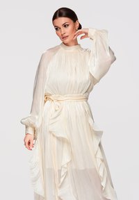 Femme en robe ivoire en satin à col montant et manches longues, ornée de volants et d'une ceinture nouée à la taille, posant avec la main près du visage sur un fond uni.