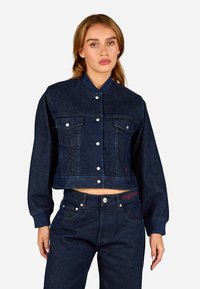 Κοντή τζιν ζακέτα, σκούρο μπλε, με κουμπιά μπροστά, δύο θησαυροφυλάκια στο στήθος και ριχτές μανσέτες. Συνδυασμένη με αντίστοιχα σκούρα τζιν παντελόνια.