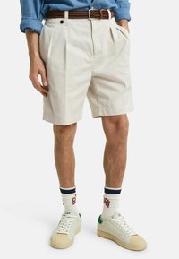 Pantalones cortos de algodón beige con pliegues, bolsillos laterales y un ajuste relajado. Combinados con zapatillas blancas y calcetines acanalados con rayas de colores.