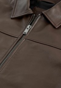 Barna bőrkabát, cipzárral, hegyes gallérral és sima textúrával. Diszkrét varrás és fémes cipzár díszíti.