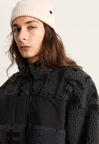 Dunkelgraue Fleecejacke mit kontrastierendem schwarzen Einsatz, hohem Kragen und durchgehendem Reißverschluss. Sie verfügt über ein strukturiertes, weiches Material für Wärme.