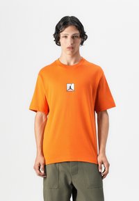 Orange bomulds T-shirt med korte ærmer og rund halsudskæring, der har en sort silhuetlogo indrammet i en hvid firkant på forsiden.
