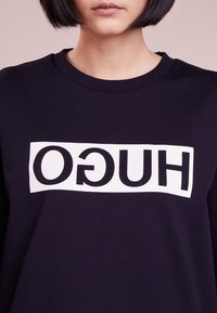 Marinblå sweatshirt med en stor vit rektangulär grafik där ordet "HUGO" står i fet svarta bokstäver. Klassisk modell med rundad halsringning. Varmt material.