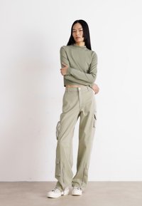 Green long-sleeve crop top paired with wide-leg cargo pants featuring side pockets and zippers. Model wearing chunky white sneakers.
