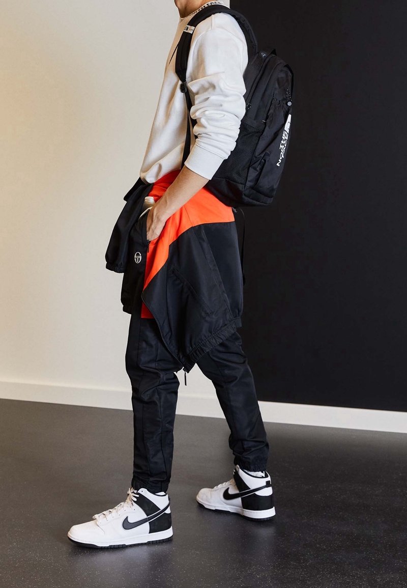 Personne portant un sweat-shirt blanc, une veste noire et orange nouée autour de la taille, un pantalon noir, des baskets Nike blanches et noires, et un sac à dos noir.