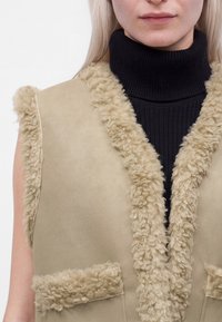 Personne blonde portant une veste beige sans manches en shearling par-dessus un pull à col roulé noir côtelé.