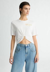 T-shirt blanc à manches courtes avec un détail noué à l'avant et le logo "LIU-JO" en doré. Associé à un jean large taille haute bleu clair.